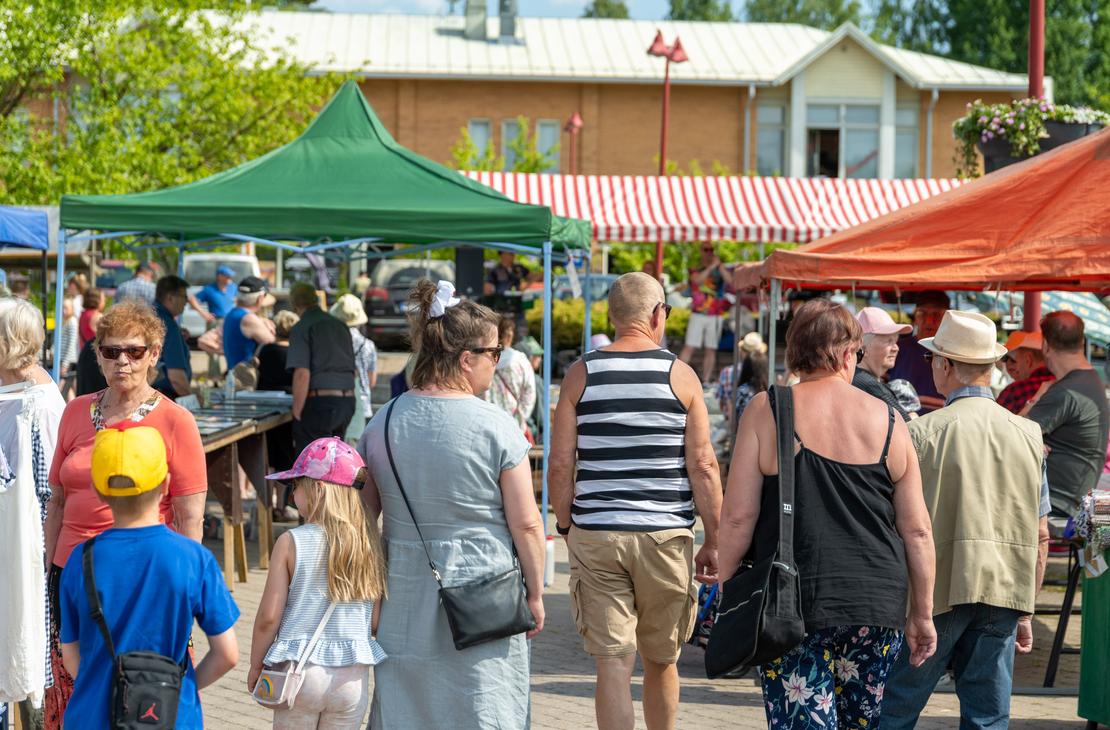 Ryhmä kesäasuisia ihmisiä kävelemässä Ilomantsissa ulkoilmatorilla vihreän ja oranssin myyntiteltan välissä kohti taustalla näkyvää esiintymislavaa, jonka yllä on puna-valkoraitainen katos.
