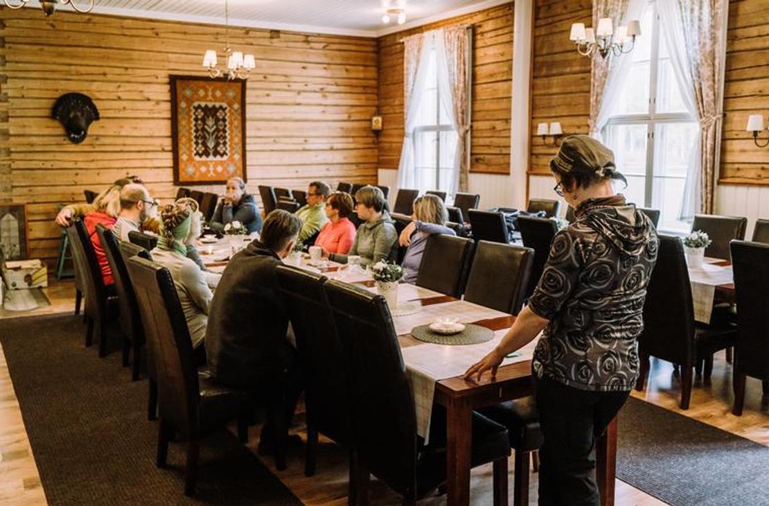 Sisäkuva Mantan Majatalon ruokasalista tai juhlatilasta. Tila on sisustettu perinteiseen tyyliin, jossa on vaaleat puupaneeliseinät ja valkoiset verhot ikkunoissa. Keskellä tilaa on pitkä pöytä, jonka ääressä on mustat tuolit ja istuu ryhmä ihmisiä. Oikealla etualalla seisoo yksi henkilö selin kameraan. Takaseinällä on kaksi koriste-esinettä, joista toinen on tumma taulu ja toinen mahdollisesti eläimen pää.