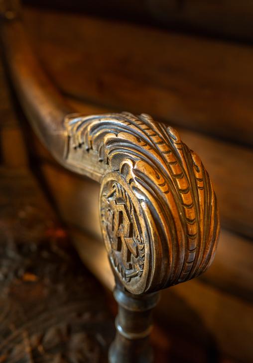 Lähikuva tumman puisen kaiteen tai tuolin käsinojan päädystä, jossa on taidokkaasti veistetty, koristeellinen kuvio. Kaiverrus on pyöreä ja kupera, muistuttaen simpukankuorta tai sulkakuviointia, ja sen keskellä on erottuva, geometrinen kuvio. Käsinoja on kiillotettu ja kiinnittyy tummaan pylvääseen. Tausta on epätarkka, mutta siinä näkyy puupintoja.
