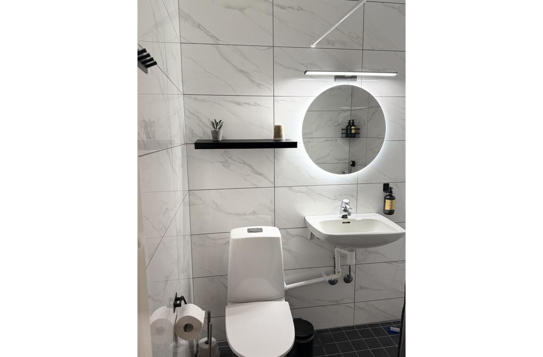 Moderni marmorilaatoitettu kylpyhuone, jossa on wc-istuin, valkoinen pesuallas sekä taustavalaistu pyöreä peili.
