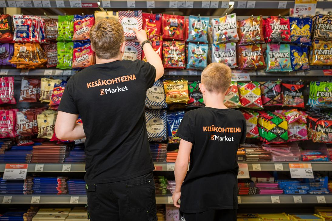 Kaksi mustiin t-paitoihin pukeutunutta henkilöä selin kuvattuna K-Market Ilomantsin karkkihyllyn edessä. Paitojen selässä on teksti ”KESÄKOHTEENA K-Market”. Vasemmanpuoleinen henkilö kurkottaa ottamaan karkkipussia ylemmältä hyllyltä, joka on täynnä värikkäitä makeispusseja, kuten TV Mix, Marianne ja Dumle.