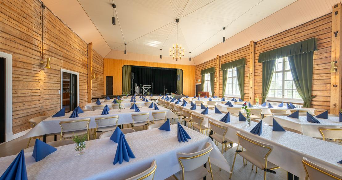 Kuva esittää Matkailukeskus Puu-Juuka Oy:n suurta juhla- tai ravintolasalia pitkien lounas- tai illallispöytien asetelmalla. Salissa on vaaleat puupaneeliseinät ja vaalea katto, josta riippuu riippuvalaisimia. Pöydät on katettu valkoisilla pöytäliinoilla ja niillä on taiteltuja sinisiä lautasliinoja. Salin päädyssä on esiintymislava keltaisella seinällä ja tummilla verhoilla. Ikkunoissa on tummanvihreät verhot.