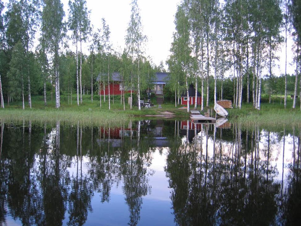 Kuva esittää Villa Blancan / LillaVilla Villan rantamaisemaa järveltä päin. Tyyni vesi etualalla heijastaa kirkkaasti taustalla olevia hoikkia koivuja ja muita puita. Puiden takaa erottuu punamultainen mökki tai sauna. Rannassa on pieni laituri ja mahdollisesti valkoinen vene tai kanootti. Maisema on rauhallinen ja kesäinen.