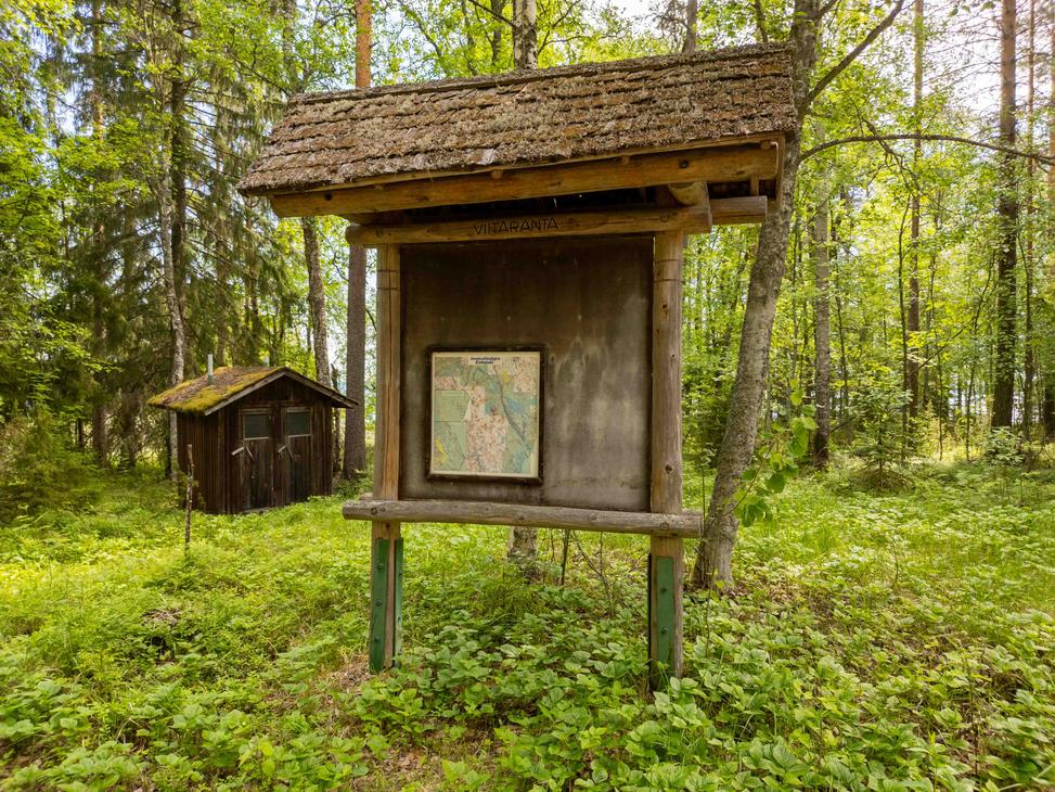 Puinen huopakattoinen opastetaulu metsän reunassa, jossa on suuri kartta ja teksti Viitaranta. Taustalla näkyy vanha tummapuinen kuivakäymälä.