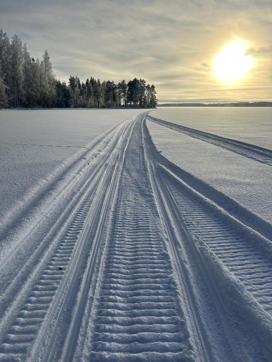 Perspektiivikuva Moottorikelkka urasta, joka kulkee lumisella järven jäällä kohti matalalla paistavaa aurinkoa ja kaukana häämöttävää metsänreunaa.