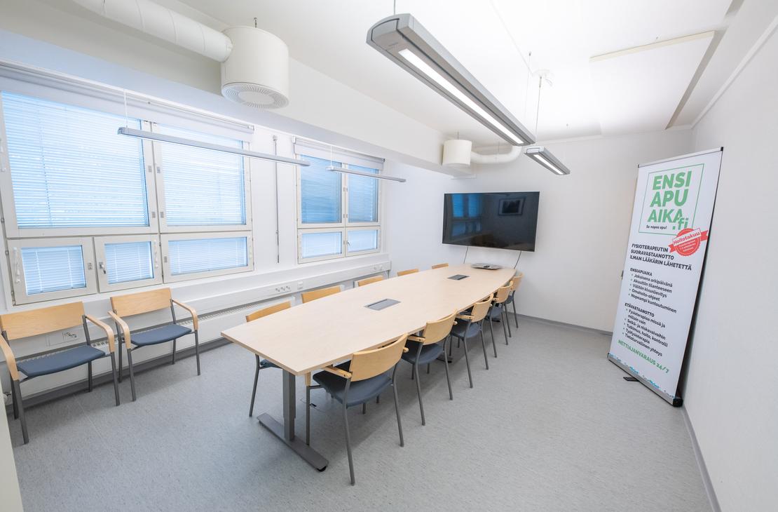 Kuva esitt&auml;&auml; Liikekulman Toimistohotellin isoa, valoisaa neuvottelutilaa tai koulutushuonetta. Huoneessa on vaalea lattia, ja keskell&auml; on suuri, vaaleapuinen neuvottelup&ouml;yt&auml;, jonka ymp&auml;rill&auml; on mustia tuoleja. P&ouml;yd&auml;n p&auml;&auml;ll&auml; on liit&auml;nt&ouml;j&auml;. Oikealla sein&auml;ll&auml; on suuri taulutelevisio ja pystyss&auml; oleva roll-up-mainos. Vasemmalla on pitk&auml; ikkunanauha, jossa on siniset s&auml;lekaihtimet, ja sein&auml;&auml; vasten on muutama ylim&auml;&auml;r&auml;inen vierastuoli.