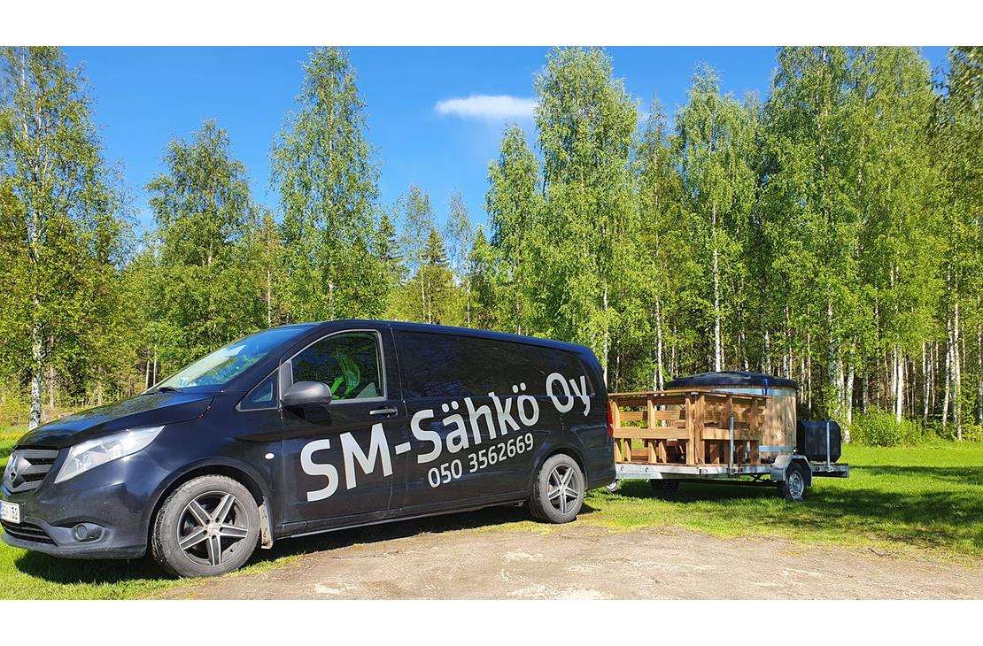 Kuva esittää SM-Sähkö Oy:n mustaa pakettiautoa, jonka kyljessä lukee valkoisella, isolla tekstillä "SM-Sähkö Oy" ja alapuolella puhelinnumero "050 3562669". Auto on pysäköity nurmialueelle, ja se vetää perässään peräkärryä, jossa on puinen, pyöreä palju tai ulkoporeamme. Taustalla on tiheä, vehreä koivumetsä aurinkoisena kesäpäivänä kirkkaansinisen taivaan alla.
