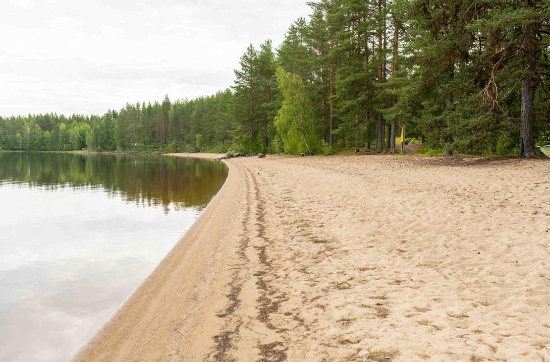 Pitkä hiekkaranta järven rannalla. Rannan vieressä kasvaa tiheä mäntymetsä, ja tyyni vedenpinta peilaa vastarannan puita.