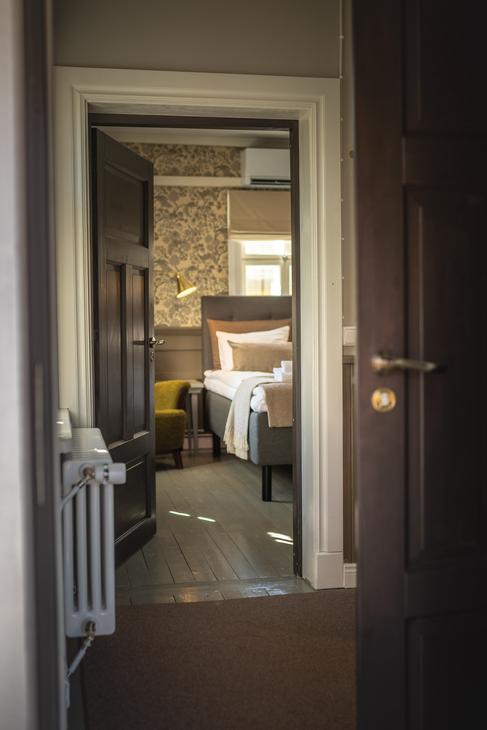 Kuva on otettu Matkailukeskus Puu-Juuka Oy:n huoneen ovelta, ja se esittää auki olevaa, tummaa puuovea, josta avautuu näkymä hotellihuoneeseen. Etualalla vasemmalla näkyy vanhanaikainen valkoinen patteri. Huoneessa, jonne ovi johtaa, on leveä sänky neutraalin sävyisillä petivaatteilla, limevihreä nojatuoli ja tumma, kuvioitu tapetti. Lattia huoneiden välillä on tummaa puuta.