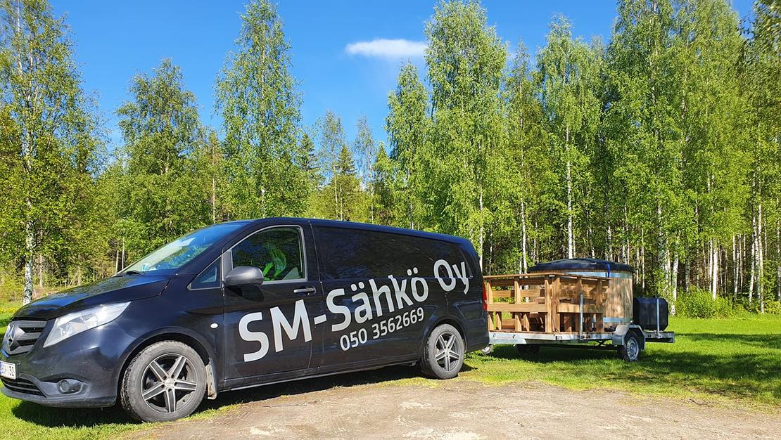 Kuva esittää SM-Sähkö Oy:n mustaa pakettiautoa, jonka kyljessä lukee valkoisella, isolla tekstillä "SM-Sähkö Oy" ja alapuolella puhelinnumero "050 3562669". Auto on pysäköity nurmialueelle, ja se vetää perässään peräkärryä, jossa on puinen, pyöreä palju tai ulkoporeamme. Taustalla on tiheä, vehreä koivumetsä aurinkoisena kesäpäivänä kirkkaansinisen taivaan alla.