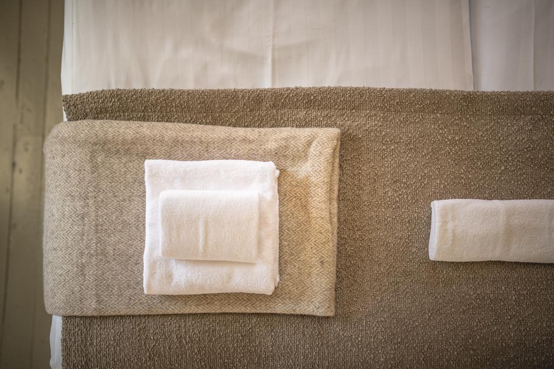 Lähikuva hotellihuoneen sängyn jalkopäästä tai päädystä Matkailukeskus Puu-Juuka Oy:ssä. Sänky on pedattu valkoisilla lakanoilla. Patjan päällä on ruskea sängynpeite tai leveä päiväpeite. Peitteen päällä on siististi taiteltu kasa valkoisia pyyhkeitä, ja oikealla on yksi erillinen pyyhe.