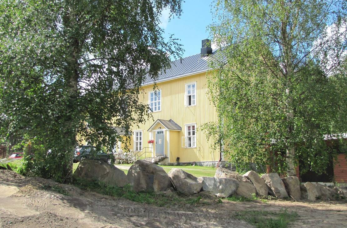 Katri Vala -kulttuurikeskuksen keltainen, kaksikerroksinen puurakennus aurinkoisena kesäpäivänä vihreiden koivujen ympäröimänä. Etualalla on hiekkamaata ja suurista luonnonkivistä koostuva matala muuri. Rakennuksessa on tumma harjakatto, valkoiset ikkunanpuitteet ja pieni katettu kuisti harmaan ulko-oven päällä.