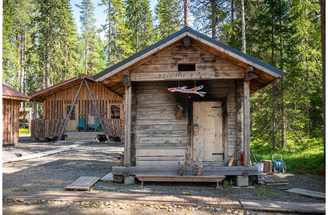 Harmaantuneesta hirrest&auml; rakennettu perinteinen sauna mets&auml;n siimeksess&auml;. Oven yl&auml;puolella on puinen kyltti, jossa lukee "MUSTA KYPY", ja terassilla on puisia koristeveistoksia.