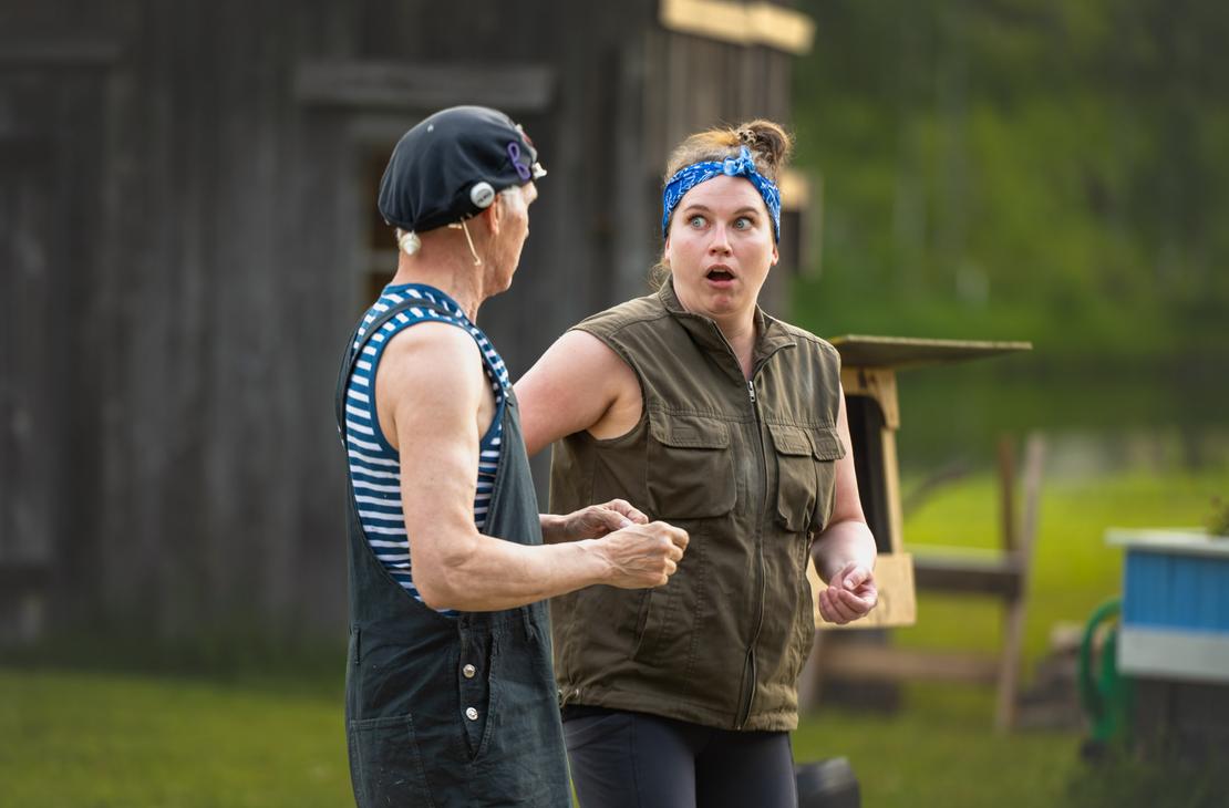 Möhkön kesäteatterin Peitetarina-näytelmän kohtaus, jossa kaksi näyttelijää esiintyy ulkonäyttämöllä vanhan puurakennuksen edustalla. Vasemmalla on raidalliseen hihattomaan paitaan, haalareihin ja mustaan päähineeseen pukeutunut mies sivuprofiilissa. Oikealla oleva nainen, jolla on sininen päähuivi ja vihreä liivi, katsoo miestä äärimmäisen hämmästyneenä suu ammollaan.