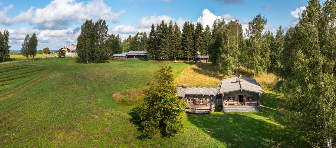 Laaja panoraamakuva, joka esittää Rajakenraalin majaa, perinteistä hirsimökkiä tai -rakennusta, joka sijaitsee avoimessa, kumpuilevassa maastossa. Rakennus on keskellä oikealla ja sen edessä on vihreä nurmikenttä. Alueella on runsaasti puita, erityisesti koivuja ja havupuita, ja taustalla avautuu maisema, jossa on lisää rakennuksia ja peltoa. Taivas on sininen ja pilvinen.