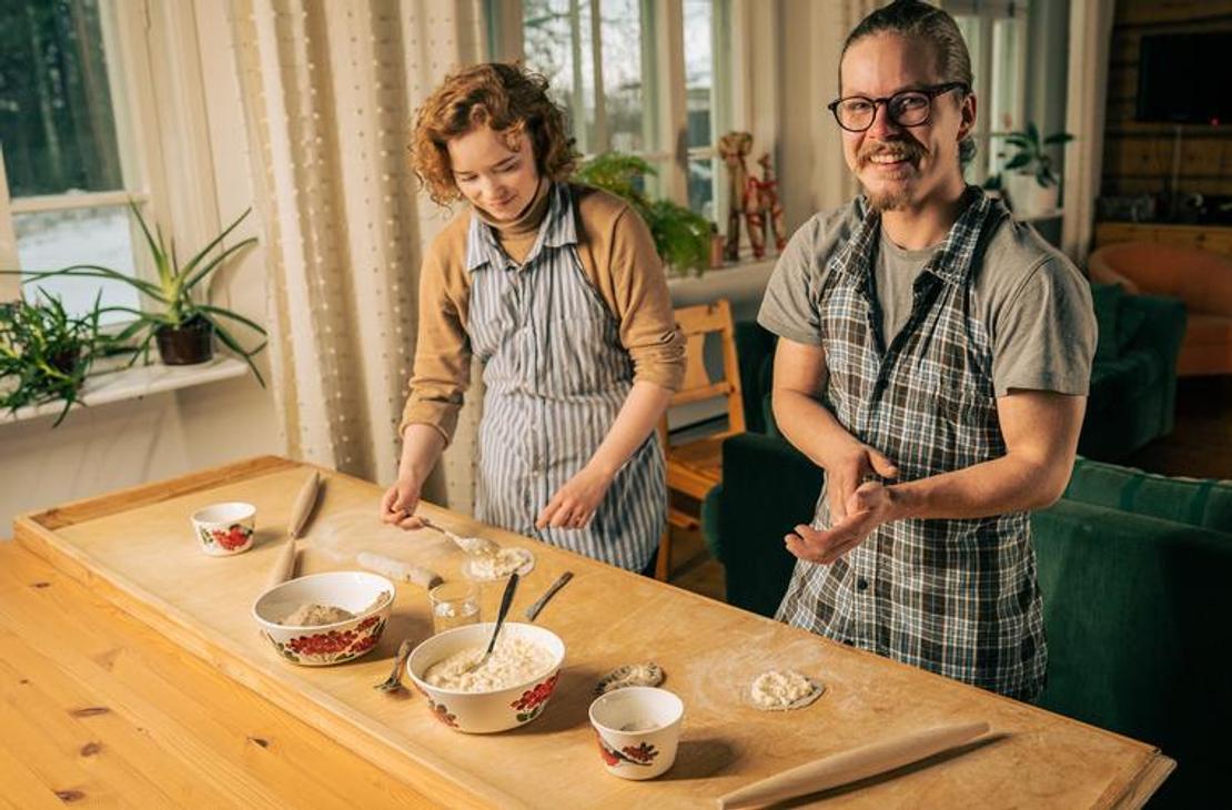 Sisäkuva, jossa kaksi aikuista, nainen ja mies, leipovat perinteisiä karjalanpiirakoita puisen pöydän ääressä. Molemmat henkilöt ovat pukeutuneet essuihin. Mies oikealla katsoo suoraan kameraan ja hymyilee. Pöydällä on taikinakulhoja ja valmiita, ohuita taikinapohjia. Tila on lämminhenkinen, ja taustalla näkyy ikkuna ja kasveja. Toiminta liittyy todennäköisesti Äksyt Ämmät -yrityksen karjalanpiirakka-työpajoihin tai -palveluihin.