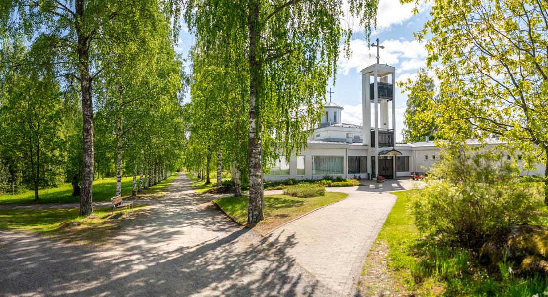 Laajakulmakuva Lintulan luostarin sisäänkäynnille aurinkoisena päivänä. Vasemmalla reunustaa vehreä koivukuja, ja oikealla laatoitettu käytävä johtaa moderniin, valkoiseen luostarirakennukseen, jossa on erillinen kellotapuli ja risti.