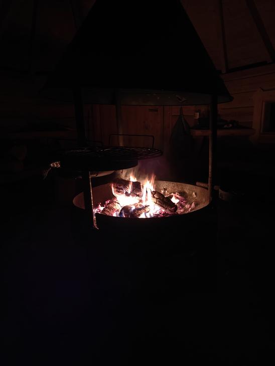 Pimeässä otettu kuva kodan tai laavun sisällä palavasta nuotiosta pyöreässä tulisijassa. Liekit valaisevat ympäröivää hämäryyttä.