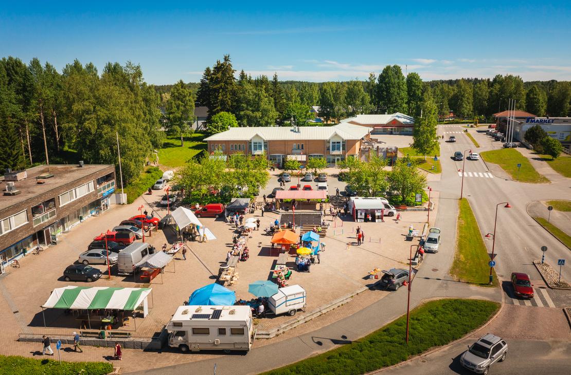 Yl&auml;viistosta kuvattu n&auml;kym&auml; aurinkoiselta torilta, jossa on useita eriv&auml;risi&auml; myyntitelttoja, ihmisi&auml; ja torimyyji&auml;. Ilomantsin torin laidalla on pys&auml;k&ouml;ityn&auml; matkailuauto ja useita henkil&ouml;autoja, ja taustalla siint&auml;&auml; S-Marketin liikerakennus sek&auml; mets&auml;&auml;.