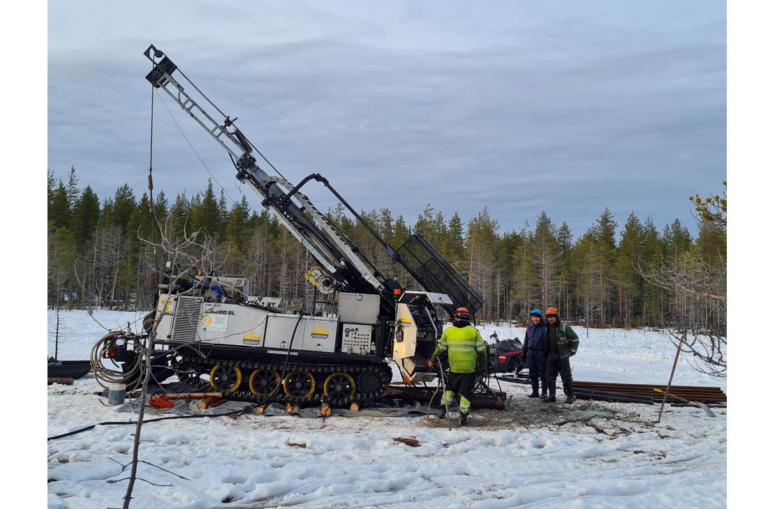 Kairaaja ja kaksi muuta henkilöä seisovat suuren telaketjuilla kulkevan kairauskoneen vieressä lumisella suolla. Taustalla on havumetsää ja pilvinen taivas.