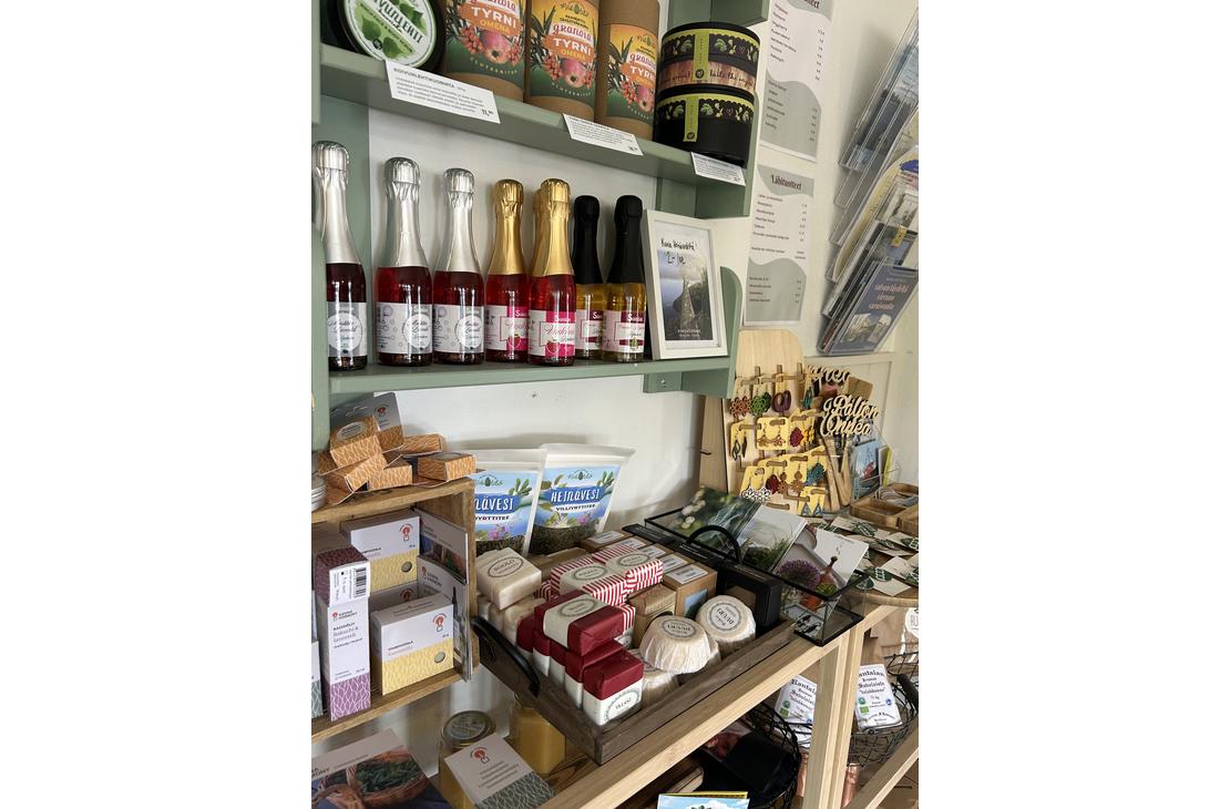 Lähikuva Satamakahvila-ravintola Kermacotan myymälähyllystä, jossa on tarjolla erilaisia paikallisia tuotteita ja matkamuistoja. Hyllyillä näkyy muun muassa pieniä kuohuviinipulloja, hilloja, paikallisia elintarvikkeita sekä käsitöitä. Tuotteet on aseteltu siististi puisiin ja vaaleanvihreisiin hyllykköihin.