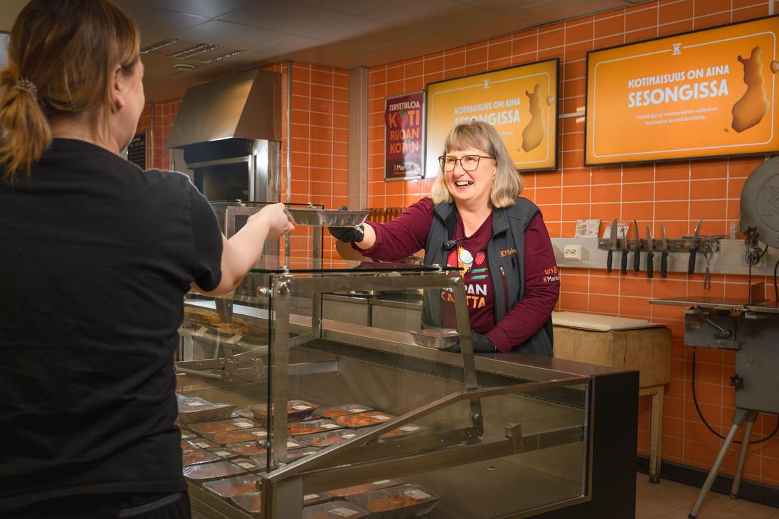 Hymyilevä K-Market Ilomantsin työntekijä ojentaa palvelutiskillä tuotetta metallivuuassa asiakkaalle lasisen suojatiskinsä yli. Taustalla on oranssi laatoitettu seinä, jossa on kyltit tekstillä ”KOTIMAISUUS ON AINA SESONGISSA”. Tiskin sisällä on esillä pakattuja tuotteita.