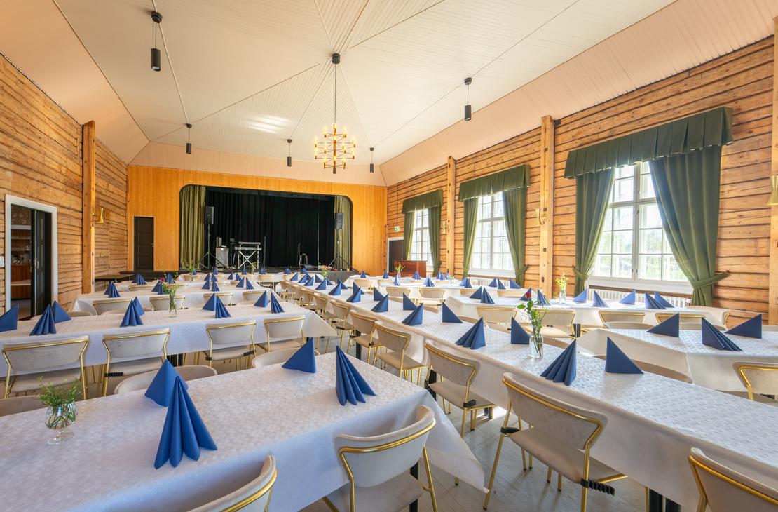 Kuva esittää Matkailukeskus Puu-Juuka Oy:n suurta juhla- tai ravintolasalia pitkien lounas- tai illallispöytien asetelmalla. Salissa on vaaleat puupaneeliseinät ja vaalea katto, josta riippuu riippuvalaisimia. Pöydät on katettu valkoisilla pöytäliinoilla ja niillä on taiteltuja sinisiä lautasliinoja. Salin päädyssä on esiintymislava keltaisella seinällä ja tummilla verhoilla. Ikkunoissa on tummanvihreät verhot.