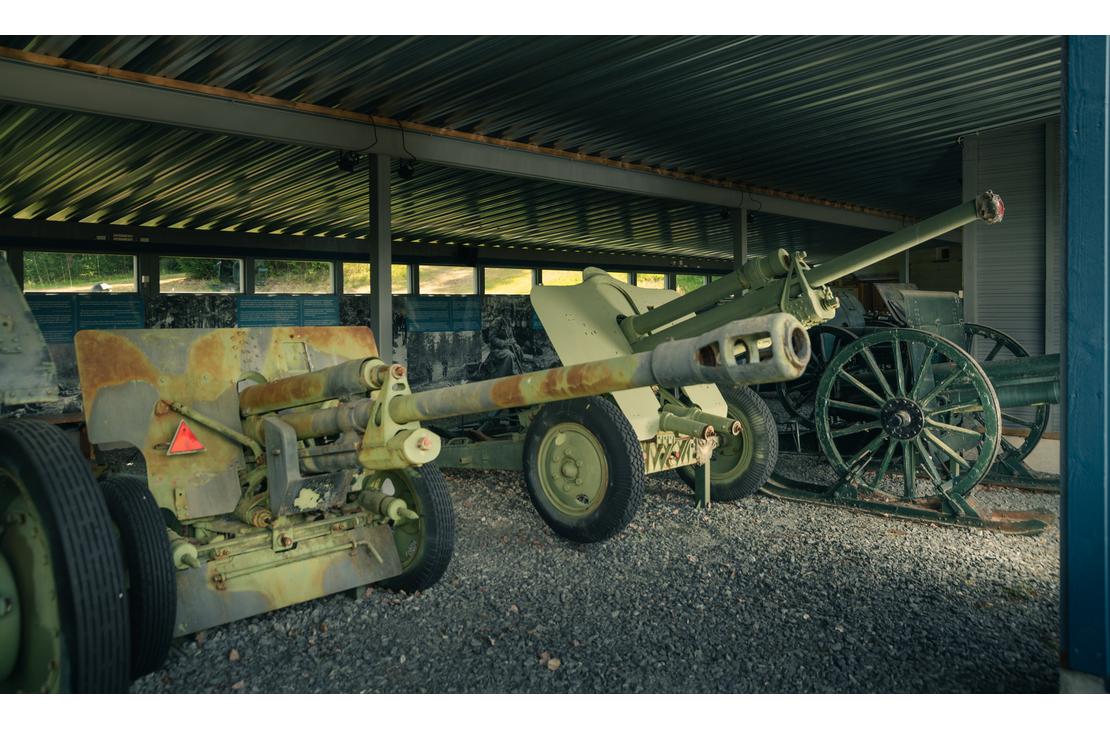 Kuva Taistelijan talon tykkikatoksesta esittää rivin historiallisia tykkejä ja aseita suojassa. Etualalla on harmaa-vihreä, maastokuvioitu panssarintorjuntatykki pyörillä. Sen takana on toinen, samankaltainen tykki, ja oikealla näkyy vanhempi, vihreä kenttätykki suurilla puupyörillä. Katoksen lattia on soraa tai hiekkaa, ja tausta on tumma.