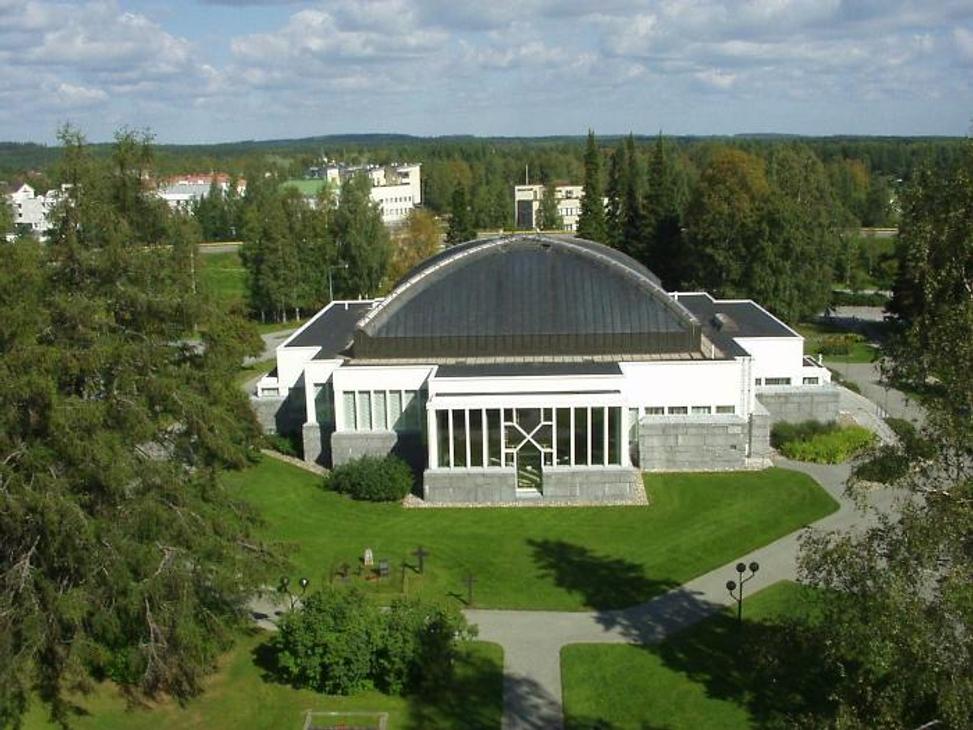 Ilmakuva Lieksan kirkosta kesällä. Valkoisessa rakennuksessa on suuri, kupolimainen tumma katto, ja sitä ympäröi vihreä nurmikko ja puusto.