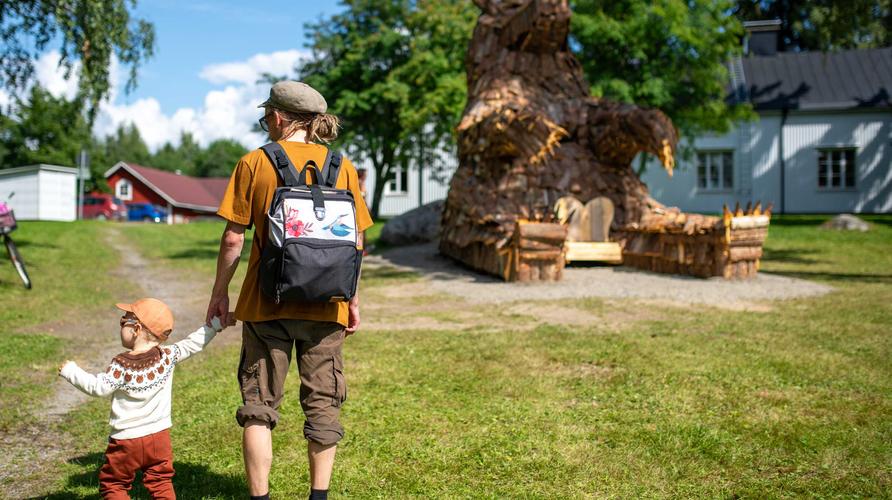Mies ja lapsi kävelevät kohti Ilokarhu -patsasta käsikädessä. Henkilöt on kuvattu takaapäin aurinkoisena päivänä.