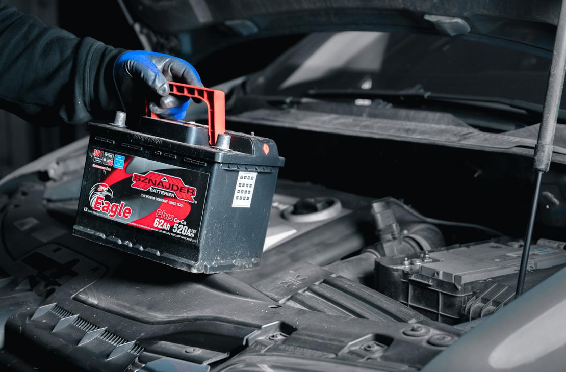 Lähikuva auton akun vaihdosta tai asennuksesta. Kuvassa nähdään käsi sinisessä hansikkaassa nostamassa tai laskemassa uutta, mustaa auton akkua, jossa on punainen kantokahva ja etiketti, harmaasävyisessä auton moottoritilassa. Konepelti on auki.