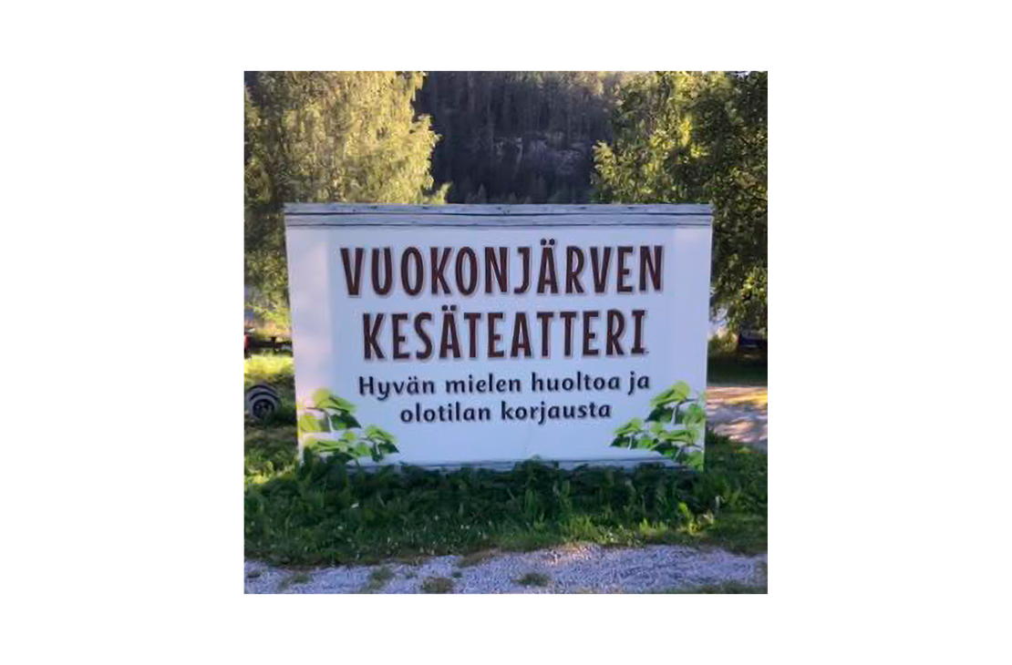 Kuva esittää Vuokonjärven Kesäteatterin opaskylttiä ulkona luonnon helmassa. Kyltti on suorakulmainen ja valkoinen. Siinä lukee tummanruskealla, isolla fontilla kaksi riviä: "VUOKONJÄRVEN" ja "KESÄTEATTERI". Sen alapuolella on pienemmällä, mustalla tekstillä tunnuslause "Hyvän mielen huoltoa ja olotilan korjausta". Kyltti on sijoitettu nurmikolle ja soraan, ja sitä kehystää ala- ja sivuosissa vihreä lehtikuviointi. Taustalla näkyy rehevää metsää.