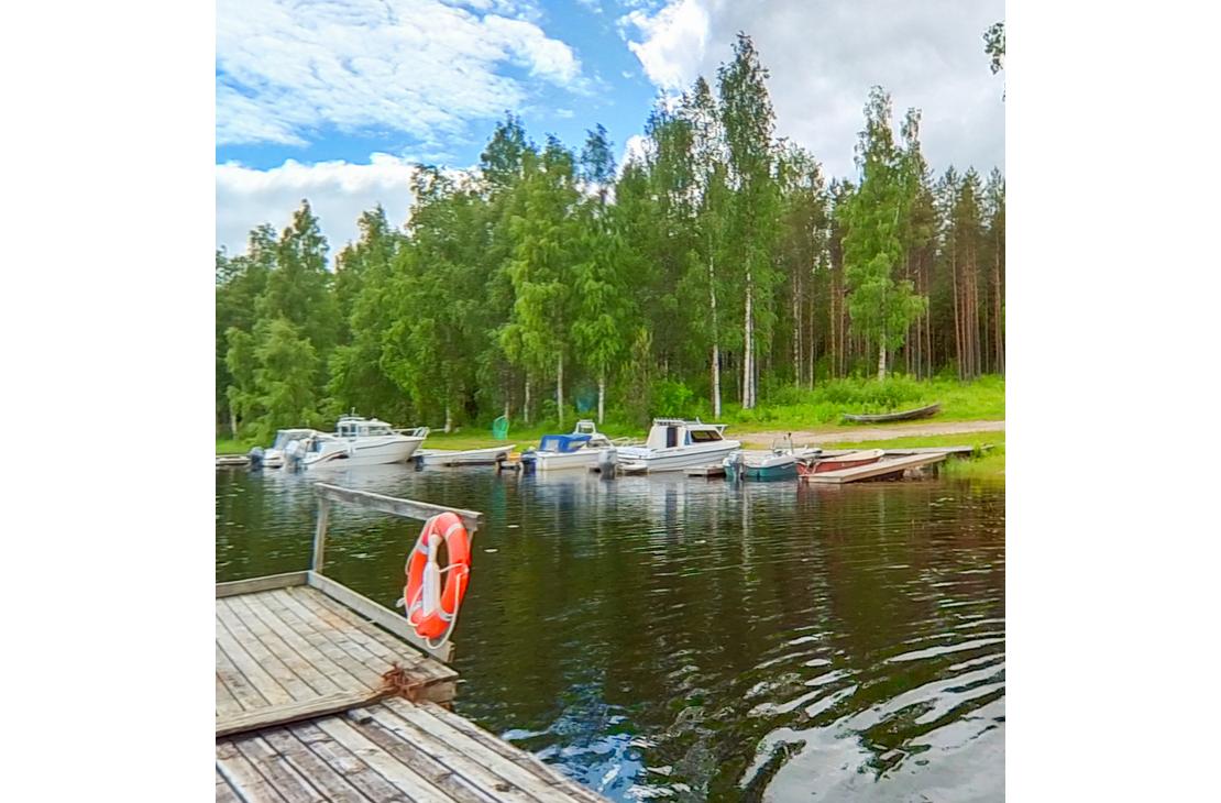 Kuva esitt&auml;&auml; pienvenesatamaa tai venelaituria aurinkoisena p&auml;iv&auml;n&auml;. Useita moottoriveneit&auml; ja pienempi&auml; soutuveneit&auml; on kiinnitetty laituriin tumman veden &auml;&auml;rell&auml;. Etualalla vasemmalla on puinen laituri, jonka kulmassa on kiinni oranssi pelastusrengas. Taustalla n&auml;kyy tihe&auml;&auml; mets&auml;&auml;, jossa on sek&auml; havupuita ett&auml; vaaleita koivuja. Taivaalla on valkoisia pilvi&auml;.