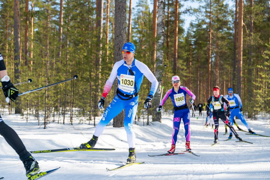 uva hiihtokilpailusta aurinkoisessa, lumisessa mets&auml;maastossa. Etualalla useita hiiht&auml;ji&auml; on liikkeess&auml;. Keskeisen&auml; hahmona on mies, jolla on sinivalkoinen, numerolla 1030 varustettu hiihtoasu, ja h&auml;n k&auml;ytt&auml;&auml; luistelutyyli&auml;. H&auml;nen takanaan on nainen pinkin- ja sinis&auml;vyisess&auml; asussa, numerolla 1060, ja muita hiiht&auml;ji&auml; kauempana. Maasto on tasaista, ja taustalla on tihe&auml;, m&auml;ntyvaltainen mets&auml;.