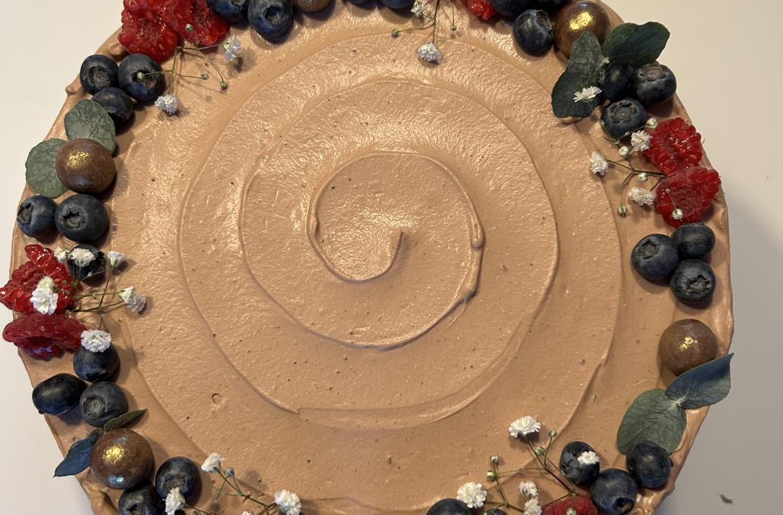 Pyöreä kakku, jonka reunoilla on runsas koristeena mustikoita, tummanpunaisia kukkia, vihreitä lehtiä ja kultaisia koristekuulia.