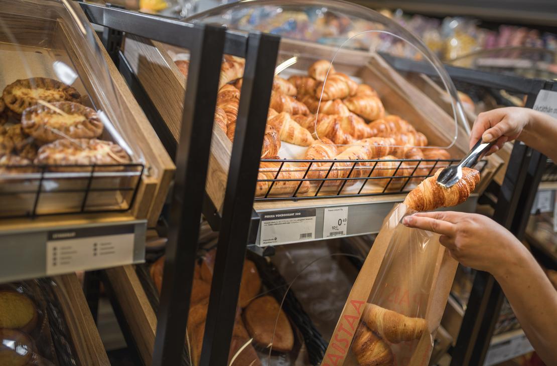 K&auml;det siirt&auml;v&auml;t metallisilla ottimilla voicroissantia paistopisteen lokerosta ikkunalliseen paperipussiin K-Market Ilomantsissa. Lokeron alla olevassa tuoteselosteessa lukee &rdquo;PIRKKA VOICROISSANT 50G&rdquo;. Vasemmalla puolella olevassa lokerossa on esill&auml; karjalanpiirakoita.