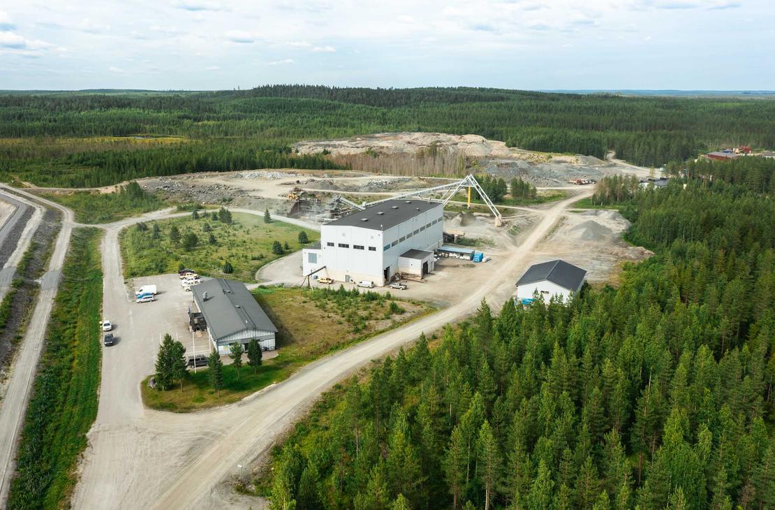 Ilmakuva Pampalon kaivokselta, joka esittää teollisuusalueen sijaintia tiheän metsän ympäröimänä. Keskellä on suuri, valkoinen jalostus- tai hallirakennus ja sen ympärillä piha-alueita sekä hiekkaisia teitä. Oikealla taustalla näkyy suuri, avoin louhosalue. Koko aluetta ympäröi tiheä, tummanvihreä havumetsä.