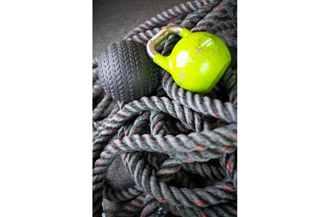 Lähikuva Kinesiocenter Kuntoilukeskuksen kuntosalilaitteista. Köysien ja painojen kasauma on etualalla: tummanharmaa harjoitusköysi (taisteluköysi), kirkkaan limevihreä kahvakuula ja tumma, kuvioitu painopallo (medicine ball). Kuva symboloi tehokasta ja monipuolista harjoittelua.