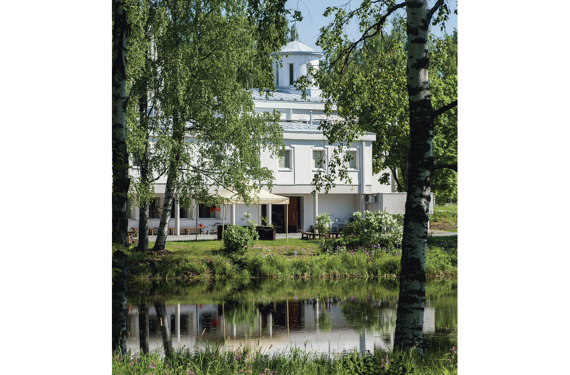 Lintulan luostarin valkoinen rakennus kuvattuna veden ääreltä kesäisenä päivänä. Etualalla on lampi, josta rakennus ja sitä kehystävät koivut heijastuvat.