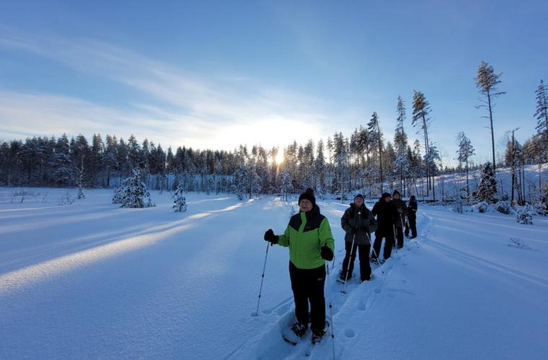 Ryhmä lumihiihdon tai lumikenkäilyn harrastajia hiihtää tai kävelee jonossa syvän, lumisen aukean yli kirkkaana talvipäivänä. Etualalla vasemmalla on yksi osallistuja kirkkaan limevihreässä takissa katsoen kameraan. Taustalla näkyy puiden ääriviivoja, jotka ovat lumen peitossa, ja aurinko paistaa matalalta horisontista luoden pitkiä varjoja.