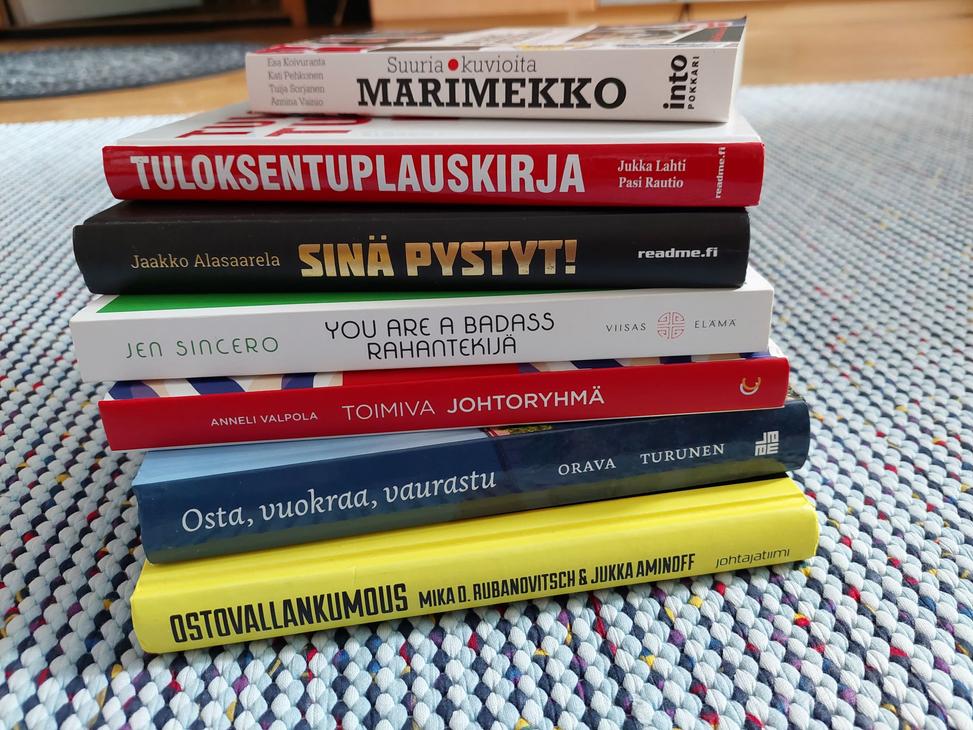 kuvassa on pino kirjoja, jotka on pinottu päällekkäin. Kirjojen kannet ovat erivärisiä ja -kuvioisia, ja niissä on erilaisia tekstejä. Pinon päällimmäinen kirja on vaaleansininen ja siinä lukee "MARIMEKKO". Pinon alimmainen kirja on keltainen ja siinä lukee "OSTA VAIN, JOS TARVITSET". Muissa kirjoissa on tekstejä kuten "TULOKSENTUPLAUSKIRJA", "SINÄ PYSTYT!", "YOU ARE A BADASS", "JOHTAJA ILMAN JOUKKOJA", "OMA RAHA, OMAT SÄÄNNÖT". Kirjat on asetettu harmaalle matolle.