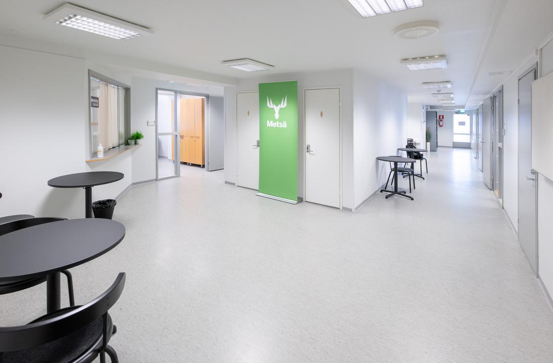 uva esittää Liikekulman Toimistohotellin modernia ja valoisaa käytävää tai aulatilaa. Käytävässä on vaalea lattia ja valkoiset seinät. Vasemmalla etualalla on pyöreitä, mustia pöytiä tuoleineen. Keskellä näkyy kirkkaan limevihreä ovi, jossa on logo (mahdollisesti yrityksen tunnus), ja taaempana oranssin ja keltaisen sävyisiä oviaukkoja. Oikealla on pitkä, valkoinen käytävä, jossa on työpöytiä.