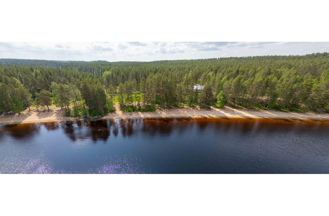 Panoraamailmakuva pitkästä hiekkarannasta, joka rajoittuu tiheään mäntymetsään. Aurinko heijastuu tumman järven pintaan ja taustalla siintää laaja metsämaisema.