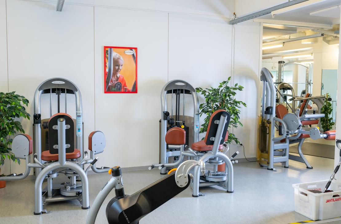 Kuva esittää osaa Kinesiocenter Kuntoilukeskuksen kuntosalitilasta, jossa on riveittäin kuntolaitteita. Laitteissa on harmaat metallirungot ja oranssinruskeat istuimet/pehmusteet. Seinät ovat valkoiset, ja niitä koristaa yksi abstrakti taulu oranssinpunaisella kehyksellä. Huoneessa on myös vihreitä huonekasveja tuomassa luonnonläheisyyttä. Lattiamateriaali on vaaleaa ja huone on hyvin valaistu.