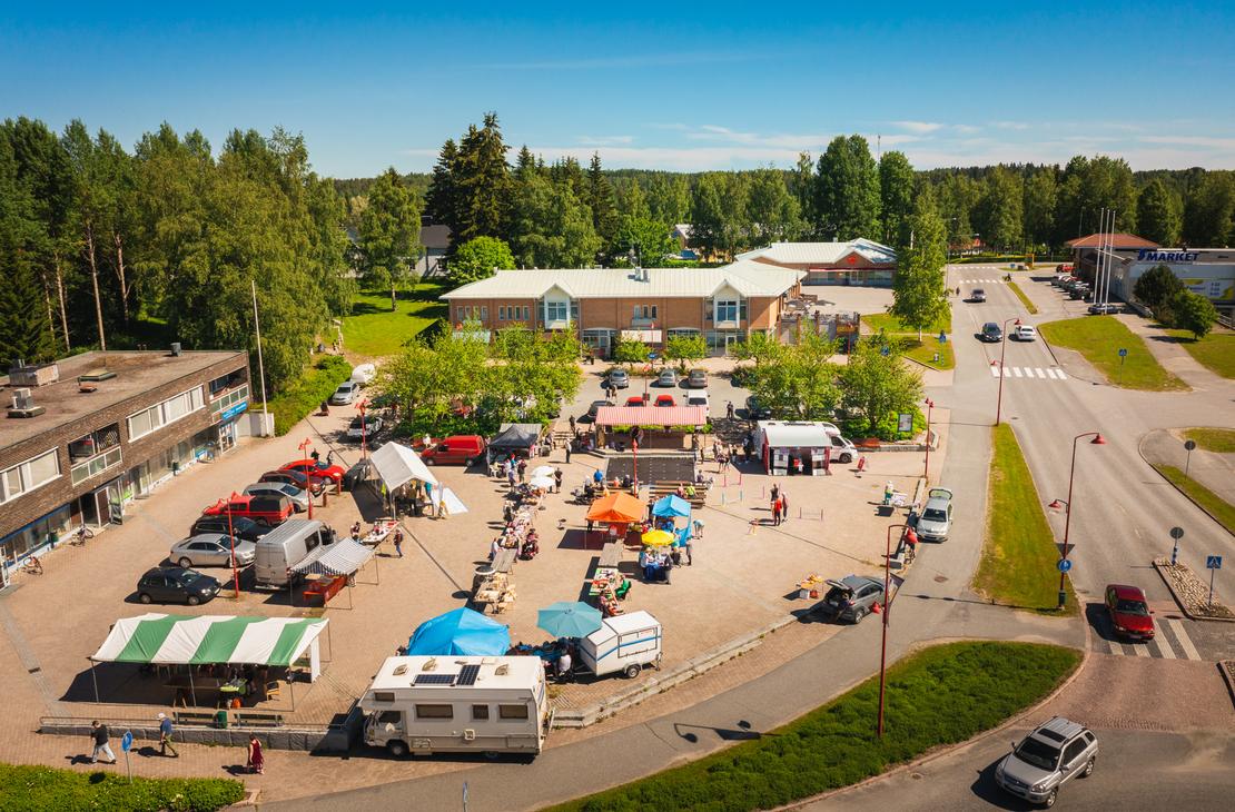 Yläviistosta kuvattu näkymä aurinkoiselta torilta, jossa on useita erivärisiä myyntitelttoja, ihmisiä ja torimyyjiä. Ilomantsin torin laidalla on pysäköitynä matkailuauto ja useita henkilöautoja, ja taustalla siintää S-Marketin liikerakennus sekä metsää.
