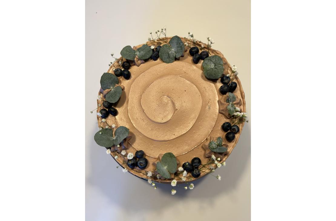 Yläviistosta otettu kuva vaaleanruskealla kuorrutteella päällystetystä kakusta, joka on koristeltu reunoilta mustikoilla, pienillä valkoisilla kukilla ja eukalyptuksen lehdillä.