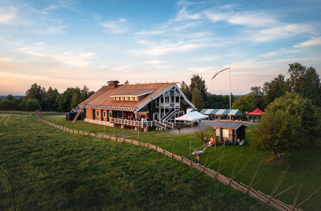 Viistosta yläkulmasta otettu kuva Parppeinpirtin hirsirakennuksesta ja sen pihasta. Etualalla on perinteinen riukuaita, joka reunustaa vihreää niittyä. Taivaanrannassa näkyy auringonlaskun värejä.