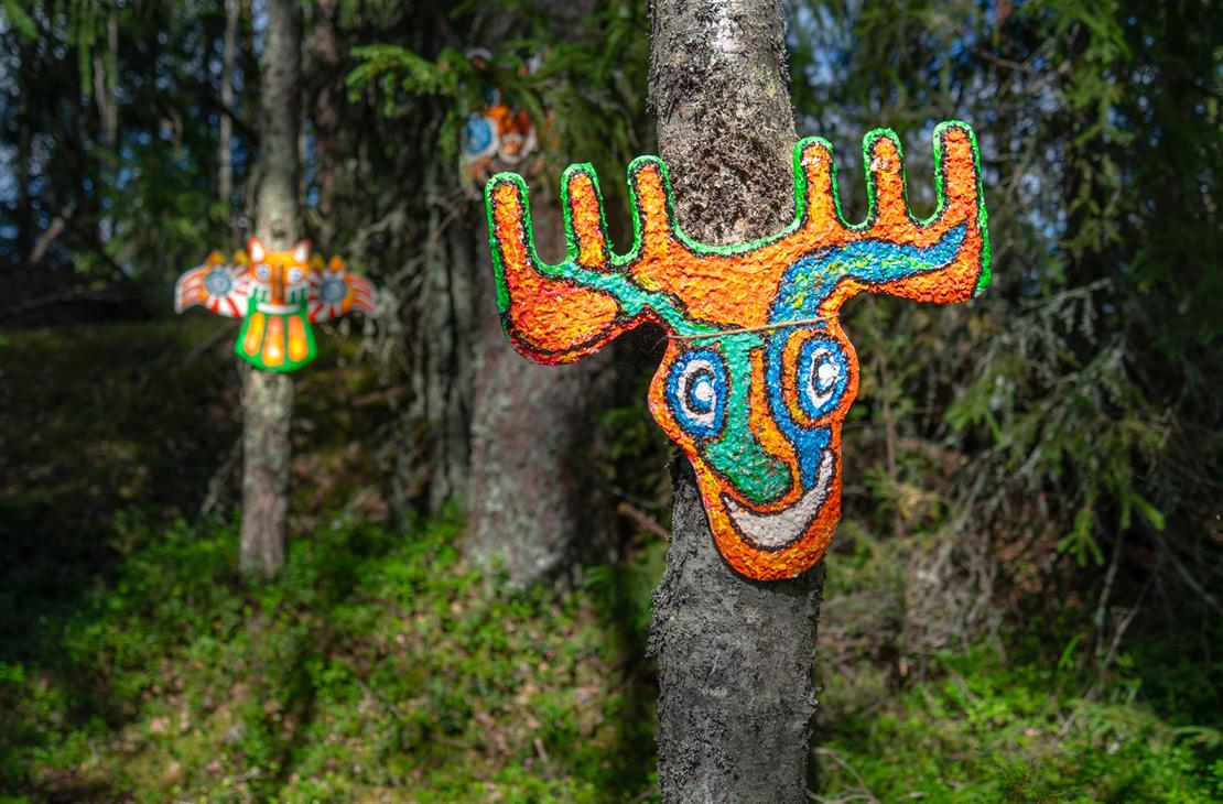 Puiden runkoihin kiinnitettyjä värikkäitä, primitiivisiä naamioita ja eläinhahmoja, kuten oranssi-vihreä hirvi- tai peura-aiheinen taideteos.