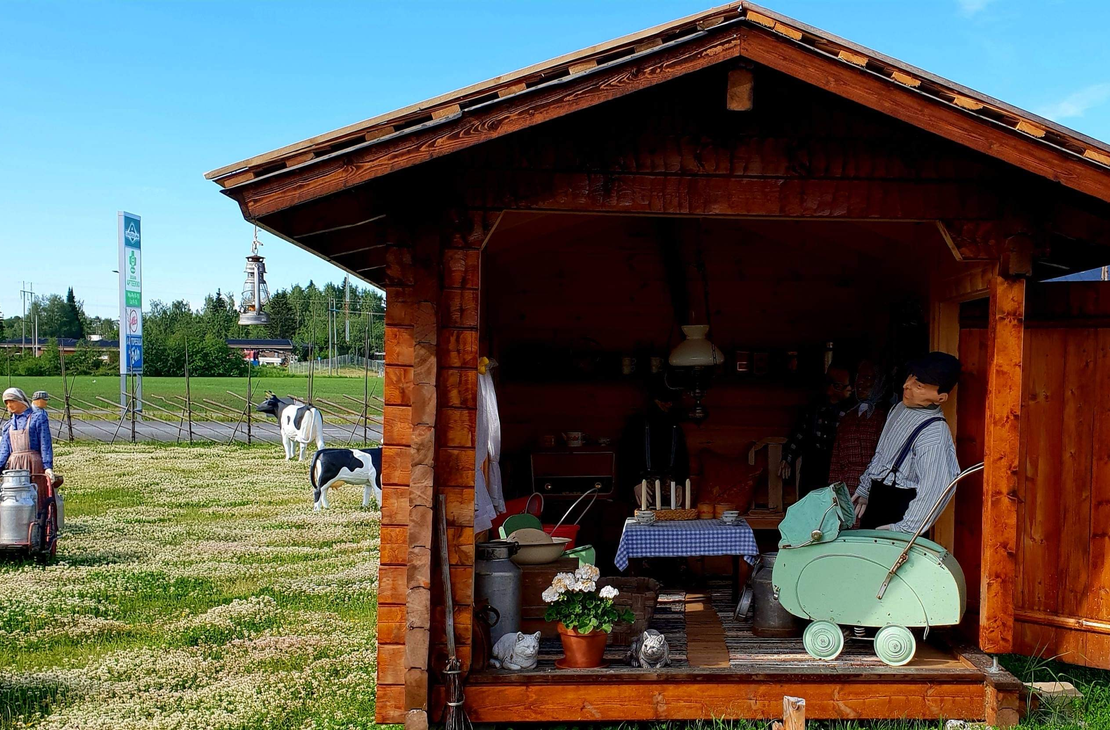 Kuva esittää Juuan Apteekin perinnepihaa aurinkoisena päivänä. Etualalla on pieni, puinen katos tai koju tummasta puusta, jossa on näytteillä vanhanaikaisia esineitä, kuten nukke tai patsas perinteisissä vaatteissa keinutuolissa sekä pöytäliinoja ja kukkia. Vasemmalla nurmialueella näkyy ihmisiä ja maalattuja lehmäpatsaita tai koristeita. Taustalla erottuu lipputanko ja kadunvarsirakennuksia.