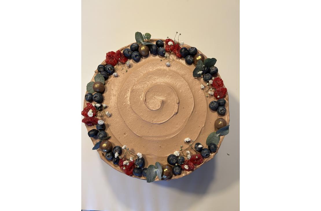 Pyöreä kakku, jonka reunoilla on runsas koristeena mustikoita, tummanpunaisia kukkia, vihreitä lehtiä ja kultaisia koristekuulia.